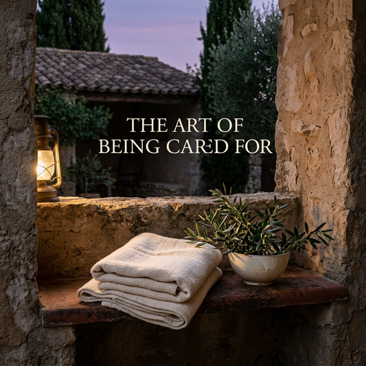 Folded ivory linen and olive branches in a ceramic bowl on a stone courtyard bench, with The Art of Being Cared For headline
